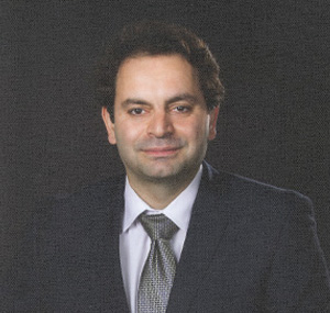 The image features a man dressed in formal attire, wearing a suit with a tie, smiling slightly at the camera. He appears to be posing for a professional portrait, likely for an identification card or similar purpose.