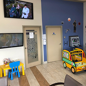 The image shows an interior space with a wall-mounted television displaying a movie scene, a blue wall with various decorations including a mural of a forest and a stylized tree, a child s play area with a colorful toy car, and a waiting room setup with seating and a door.