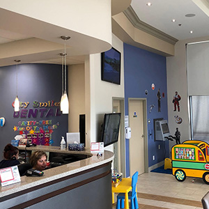 The image shows an interior view of a dental office with a playful and child-friendly atmosphere, featuring a cartoon character on a sign, a toy truck, and a waiting area with children s artwork on display.